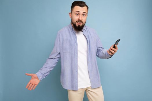 Upset handsome young brunette unshaven man with beard wearing stylish white t-shirt and blue shirt isolated over blue background with empty space holding in hand and using phone messaging sms looking at camera and having troubles.