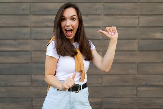 Portrait of young happy positive beautiful brunette woman with sincere emotions wearing stylish outfit standing in the street near the wall and pointing at herself. Lifestyle concept.