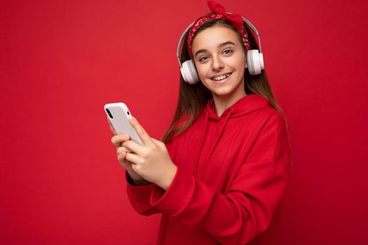 Closeup portrait photo of beautiful positive smiling brunet female teenager wearing red hoodie isolated on red background holding and using smartphone communicating online wearing white wireless headphones listening to cool music looking at camera.