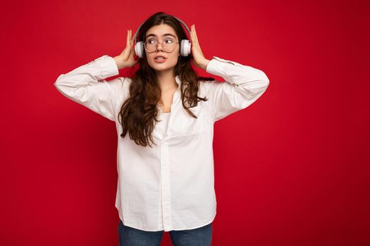 Attractive young brunette female person wearing white shirt and optical glasses isolated over red background wearing white wireless bluetooth earphones listening to music and looking to the side thinking about smth keeping hands near head.