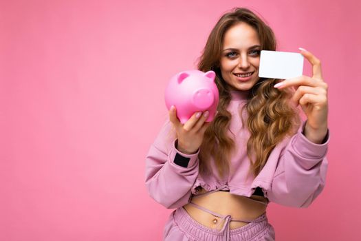 Shot of happy positive smiling young winsome attractive woman with wavy long blonde hair with sincere emotions wearing stylish pink hoodie isolated over pink background with copy space, holds pink pig penny bank for money and credit card. Savings concept.