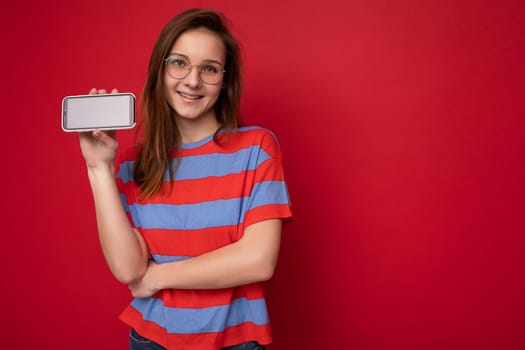 Smiling attractive positive good looking young woman wearing optical glasses and casual stylish outfit poising isolated on background with empty space holding in hand smartphone and showing phone empty screen looking at camera.