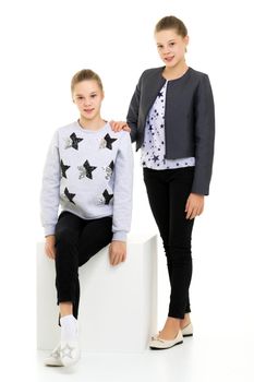 Pretty Sisters Standing Leaning on Cube with one Knee and Looking at Camera, Full Length Portrait of Two Teen Girls Wearing Trendy Stylish Clothes Posing Together in Studio Against White Background