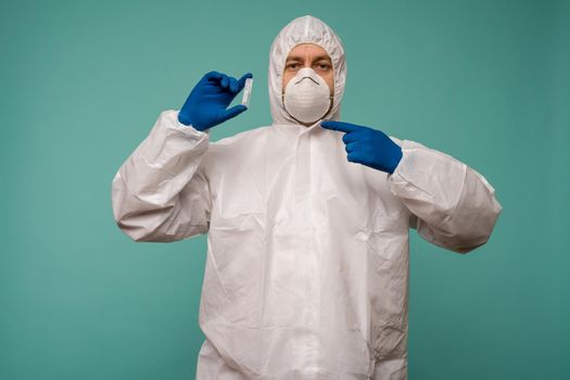 A male doctor in protective overalls and a mask pointing at ampoule with a vaccine in his hands. Coronovirus protection concept in China. - image