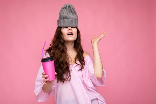 Pretty young positive brunette curly woman wearing pink shirt and grey hat isolated over pink background holding paper coffee cup for mockup drinking and having fun.