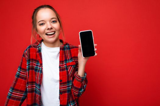 Photo of beautiful cute positive young blonde woman standing isolated over red wall wearing casual red shirt and white t-shirt showing mobile phone with empty screen for mockup looking at camera. copy space