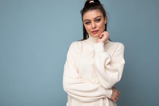 Young brunette woman with wearing white casual sweater isolated over blue background looking confident at the camera with crossed arms and hand raised on chin. Thinking positive.