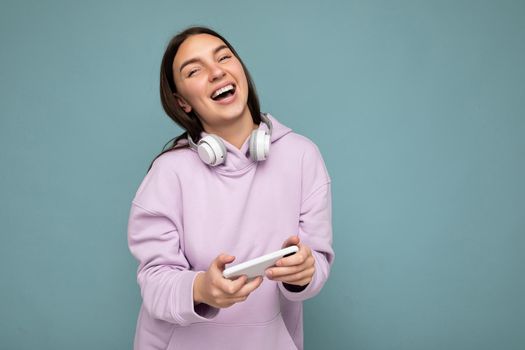 Beautiful happy smiling young brunette female person wearing light purple hoodie isolated over blue background wall usine smartphone wearing white bluetooth headsets listening to cool music and looking at camera. empty space