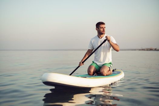 Young man in t-shirt and shorts floating on SUP board at sea
