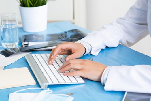 Close-up of female doctor hands typing at computer keyboard. Physician in medical uniform working at desk. Examination and diagnosis in hospital. Practitioner workplace with computer keyboard.
