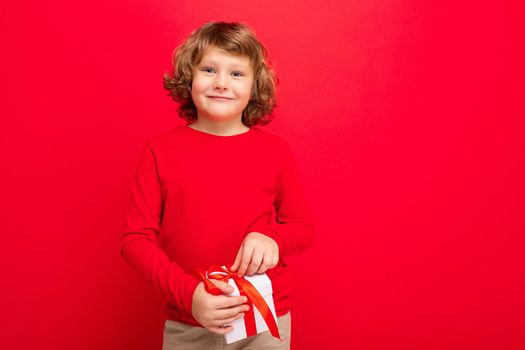 Positive smiling blonde curly boy isolated over red background wall wearing red sweater holding gift box and looking at camera. Copy space