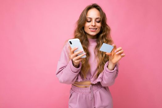 Beautiful smiling positive young blonde curly woman wearing pink clothes isolated over pink background using mobile phone paying online shopping through credit card looking at smartphone display.