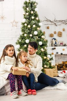 Merry Christmas and Happy Holidays. Cheerful mom and her cute daughters girls exchanging gifts. Parent and two little children having fun and playing together near Christmas tree indoors