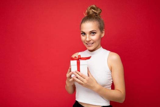 Shot of beautiful positive smiling young blonde woman isolated over red background wall wearing white top holding gift box and looking at camera. Copy space, mockup