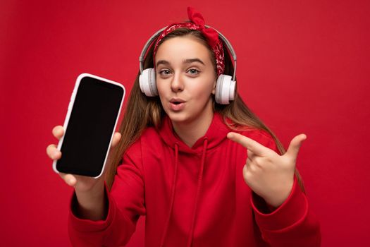 Attractive joyful amazed brunette girl wearing red hoodie isolated on red background holding and showing smartphone with empty display for cutout wearing white wireless headphones listening to funny music looking at camera and pointing finger at device.