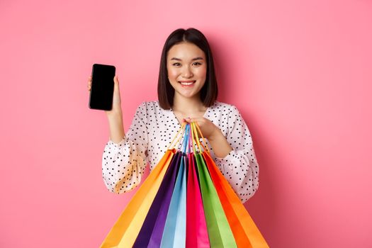 Attractive asian woman showing smartphone app and shopping bags, buying online via application, standing over pink background.