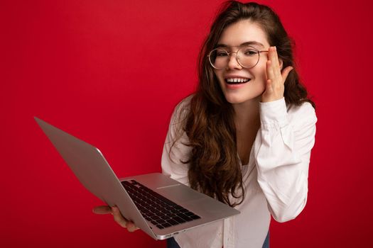 Side profile Photo shot of beautiful smiling brunet curly young woman holding computer laptop wearing glasses white shirt looking at netbook display isolated over red wall background.