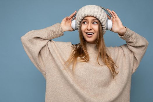 Photo of beautiful sexy happy smiling young blonde woman wearing beige winter sweater and hat isolated over blue background wearing white wireless bluetooth headphones listening to cool music dancing and looking to the side.