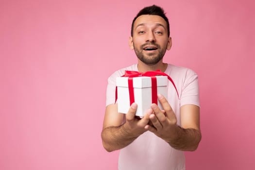 Photo shot of handsome positive happy kind brunette young unshaven man with beard isolated over pink background wall wearing casual pink t-shirt holding white gift box with red ribbon and looking at camera. Copy space, mockup
