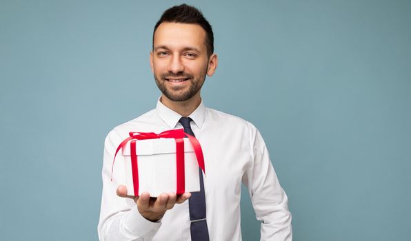 Photo shot of handsome happy smiling brunet young unshaven man isolated over blue background wall wearing white shirt and tie holding white gift box with red ribbon and looking at camera.