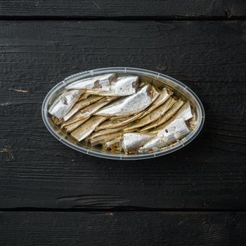 Canned Anchovy set, in plastic container, on black wooden table background, top view flat lay, square format