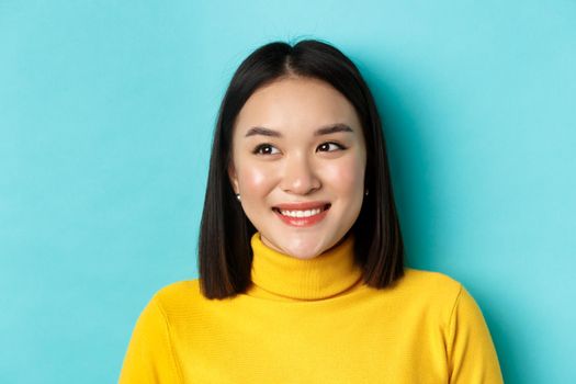 Beauty and makeup concept. Dreamy asian girl with perfect healthy skin, looking left and smiling happy, standing over blue background.