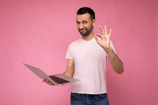 Handsome funny brunet man holding laptop computer and showing gesture ok looking at camera in t-shirt on isolated pink background.