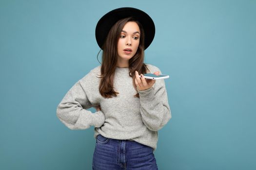 young asking dissatisfied brunette woman speaking wearing black hat and grey sweater holding smartphone looking to the side isolated on background.