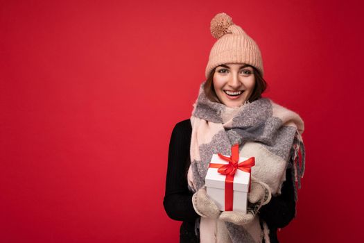 Beautiful happy young brunette woman isolated over colourful background wall wearing stylish casual clothes holding gift box and looking at camera. Free space