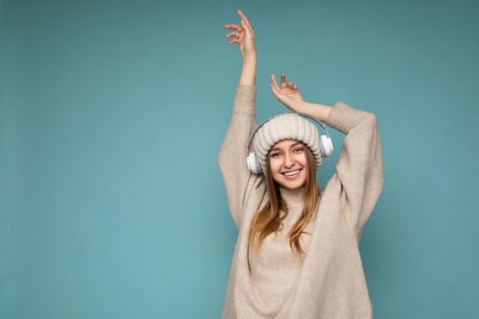 Attractive positive smiling young blonde woman wearing beige winter sweater and hat isolated over blue background wall wearing white wireless bluetooth earphones listening to music having fun and looking at camera. free space