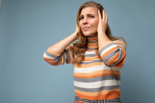 Photo of young dissatisfied attractive blonde curly woman with sincere emotions wearing casual striped pullover isolated on blue background with copy space and covering ears trying not to hear.