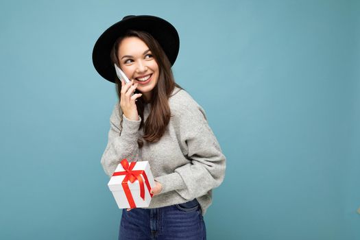 Photo of beautiful happy positive joyful young brunette woman isolated over blue background wall wearing black hat and grey sweater holding gift box talking on mobile phone and looking to the side. Copy space, mockup