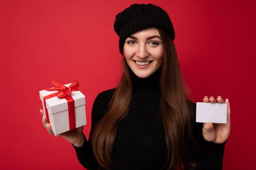 Closeup portrait Attractive positive young brunette woman wearing black sweater and hat isolated on red background holding credit card and gift box looking at camera.