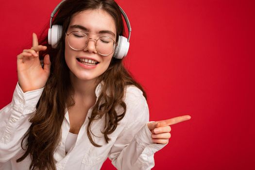Closeup photo of attractive positive smiling young brunet woman wearing white shirt and optical glasses isolated over red background wearing white wireless bluetooth headsets listening to music and moving having fun. free space