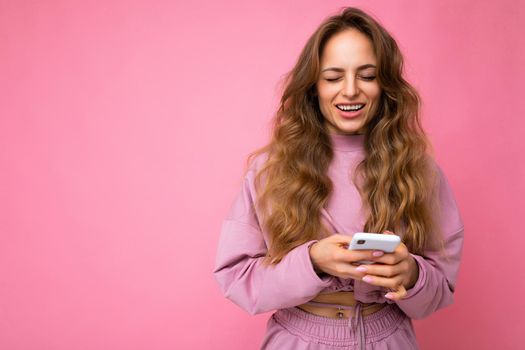 Photo shot of winling attractive positive good looking young woman wearing casual stylish outfit poising isolated on background with empty space holding in hand and using mobile phone messaging sms looking at camera.