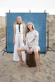 beauty portrait outdoors on sand in front of blue background, young pretty twins with long blond hair posing in sand quarry in elegant white, beige clothes. identical caucasian sisters pose in hats