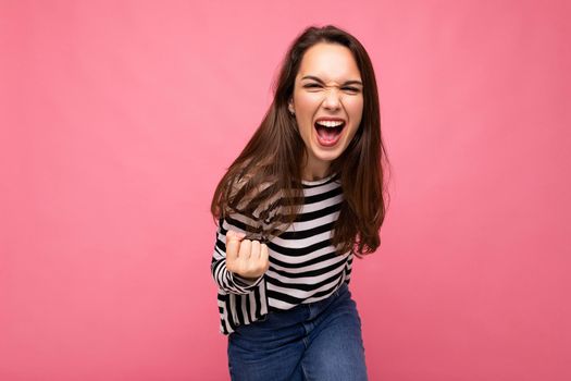 Portrait of young positive happy beautiful brunet female person with sincere emotions wearing casual striped pullover isolated on pink background with empty space and celebrating winning shouting yes.