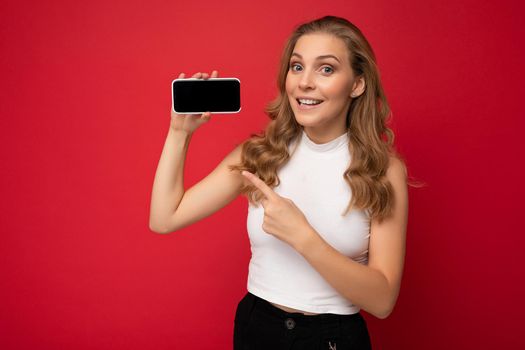 Photo of beautiful smiling young woman good looking wearing casual stylish outfit standing isolated on background with copy space holding smartphone showing phone in hand with empty screen display for mockup pointing at gadjet looking at camera.