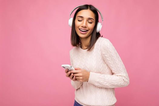 Photo shot of beautiful joyful smiling young female person wearing stylish casual outfit isolated over colorful background wall wearing white bluetooth wireless earphones and listening to music and using mobile phone with close eyes. copy space