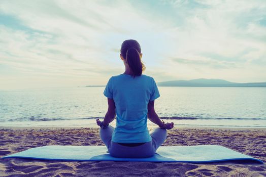 Young woman meditating in pose of lotus on beach near the sea in summer in the morning, rear view