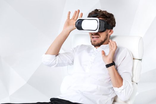 Excited young man is getting experience using VR-headset glasses of virtual reality gesticulating with his hands. A young man sits on a comfortable armchair in a room with white walls