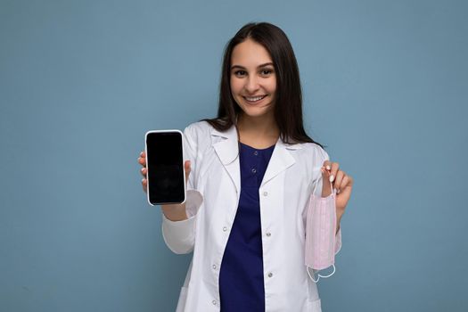 Photo of beautiful smiling young woman good looking wearing casual stylish outfit standing isolated on background with copy space holding smartphone showing phone in hand with empty screen display for mockup looking at camera.