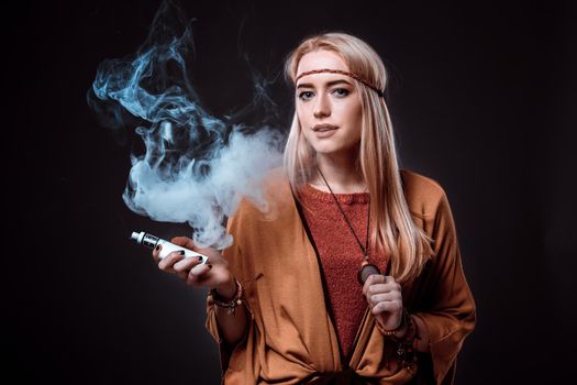 Young woman in the Boho style blowing smoke. The blonde on a dark background. Young woman looking into the camera