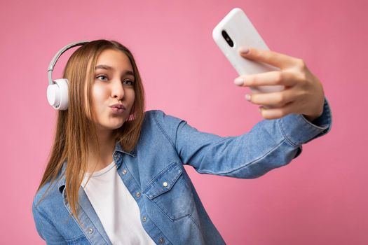 Sexy attractive happy smiling young blonde woman wearing blue jean stylish shirt and casual white t-shirt isolated over pink background wearing white bluetooth wireless headphones and listening to good music and using mobile phone having video call communication looking at gadjet and giving kiss.