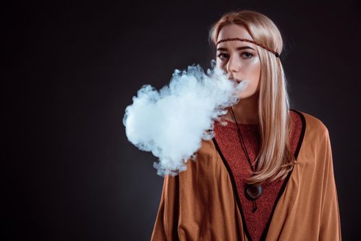 Young woman in the Boho style blowing smoke. The blonde on a dark background. Young woman looking into the camera