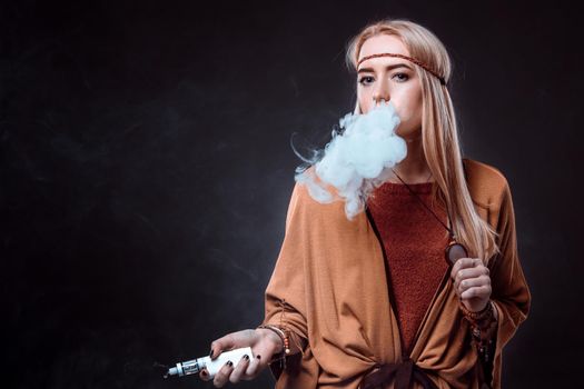 Young woman in the Boho style blowing smoke. The blonde on a dark background. Young woman looking into the camera