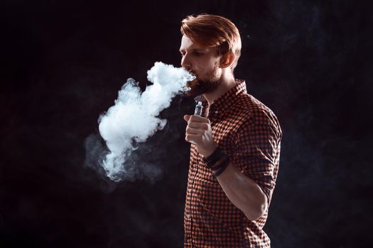 young man wearing a plaid shirt smokes an electronic cigarette on a black background