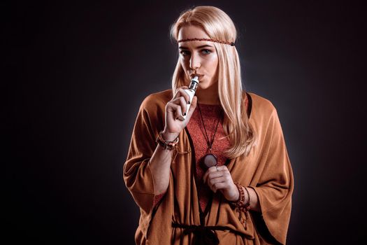 Young woman in the Boho style blowing smoke. The blonde on a dark background. Young woman looking into the camera