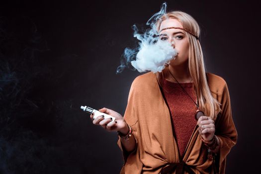 Young woman in the Boho style blowing smoke. The blonde on a dark background. Young woman looking into the camera