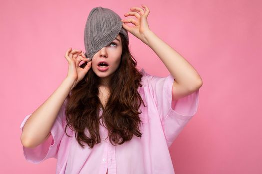 Photo shot of charming crazy emotional young brunette woman standing isolated over pink background wall wearing pink shirt and gray hat and having fun.